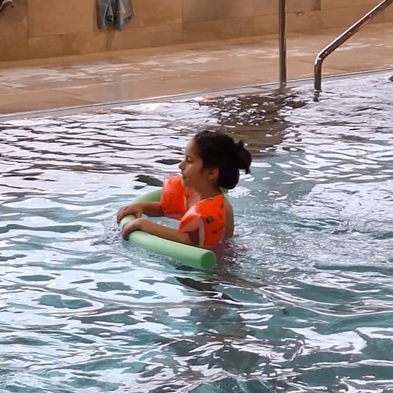Ein junges Mädchen in einem orangefarbenen Schwimmweste treibt auf dem Rücken im Pool und hält einen grünen Schwimmhilfe.