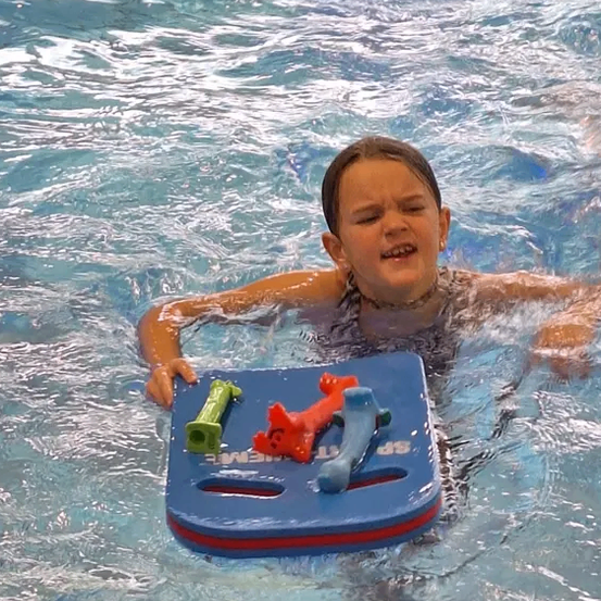 Ein kleiner Junge schwimmt auf einem blauen Brett im Schwimmbad mit drei Spielzeugen. Er lächelt.