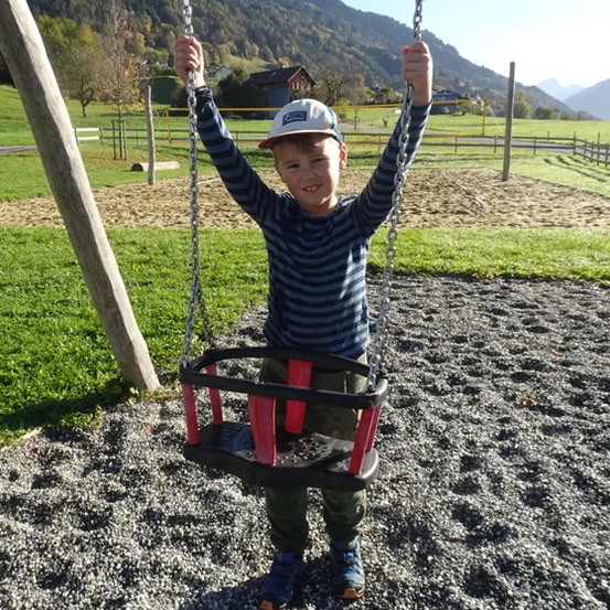 Ein junger Junge steht auf einem roten Schaukel, wobei er die Ketten festhält. Er lächelt, trägt einen Hut und Turnschuhe. Hinter ihm befindet sich ein Grasbereich mit einem Zaun und Bergen in der Ferne.