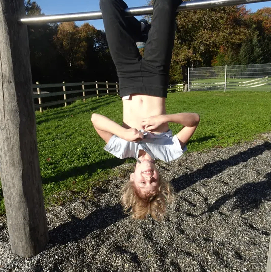 Ein Kind hängt kopfüber an einer Stange auf einem Spielplatz. Der Boden ist aus Kies, und im Hintergrund befindet sich ein Zaun.