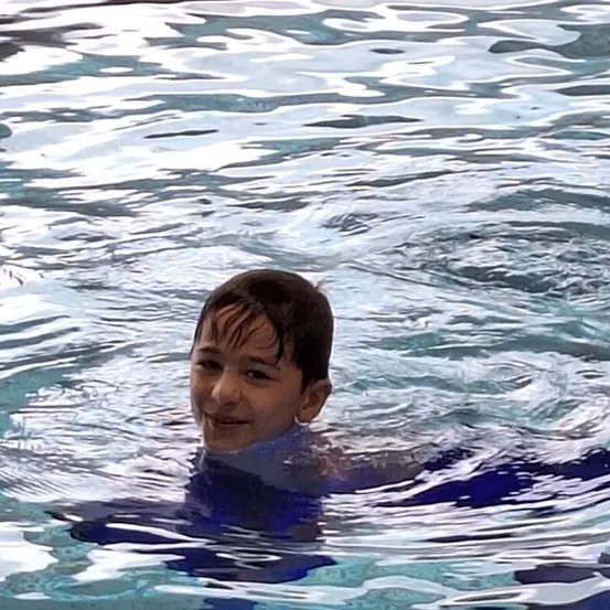 Ein junger Junge schwimmt mit fröhlicher Miene in einem Pool und trägt einen blauen Badeanzug. Das Wasser ist ruhig mit kleinen Wellen.
