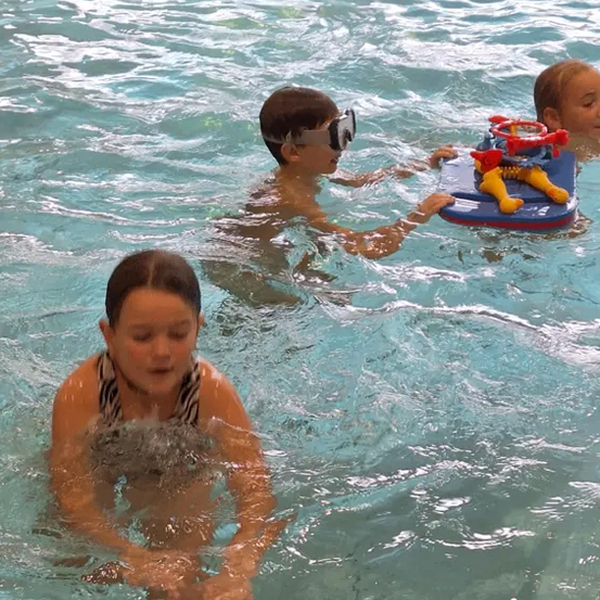 Drei Kinder schwimmen in einem Pool. Ein Junge mit Schwimmbrille hält ein Spielzeugboot. Ein anderes Kind lächelt in die Kamera. Das Poolwasser ist klar.