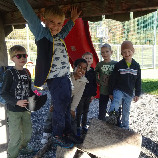 Sechs junge Jungen stehen auf einem Spielplatz mit einer roten Rutsche im Hintergrund. Ein Junge steht auf einem Baumstamm, andere lächeln. Sie tragen Jacken und Turnschuhe.