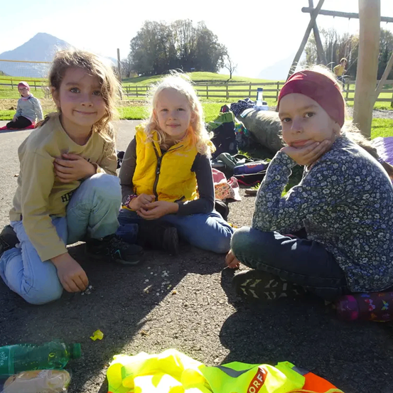 Drei junge Mädchen sitzen auf dem Boden, wahrscheinlich in einem Park, und lächeln in die Kamera. Ein gelber Sicherheitsweste und verschiedene Gegenstände sind um sie herum verstreut. Im Hintergrund ist ein Rasenbereich mit einem Zaun und einem Spielplatz zu sehen.