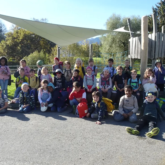 Eine Gruppe von Kindern posiert für ein Foto auf einem Spielplatz mit einem großen Vordach, Bäumen und Bergen im Hintergrund.