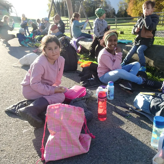 Mehrere Kinder sitzen auf dem Boden in einem Park. Zwei Mädchen lächeln. Es gibt Rucksäcke, Taschen und Wasserflaschen um sie herum. Im Hintergrund sitzen mehr Kinder auf einem Holzstamm.