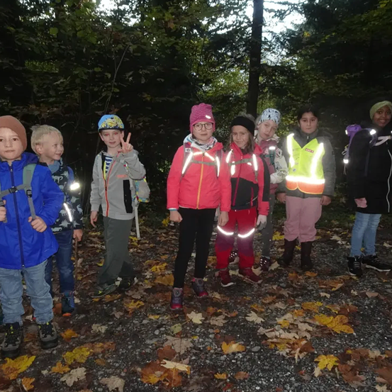 Eine Gruppe von Kindern, alle mit Sicherheitswesten, steht in einem Wald mit Blättern auf dem Boden. Einige tragen Rucksäcke.
