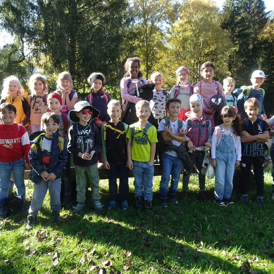 Eine Gruppe von Kindern in einem Park mit hohen Bäumen im Hintergrund, einige tragen Rucksäcke und Hüte.
