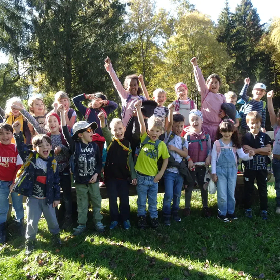 Eine Gruppe von Kindern und Jugendlichen posiert für ein Foto in einem Park mit Bäumen im Hintergrund. Einige von ihnen heben ihre Hände.