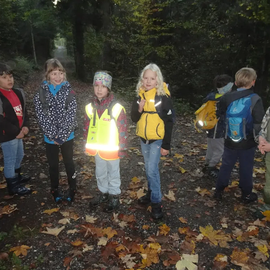 Eine Gruppe von Kindern steht in einem Wald, trägt Jacken und Rucksäcke. Blätter sind auf dem Boden verstreut. Ein Kind trägt eine reflektierende Weste.