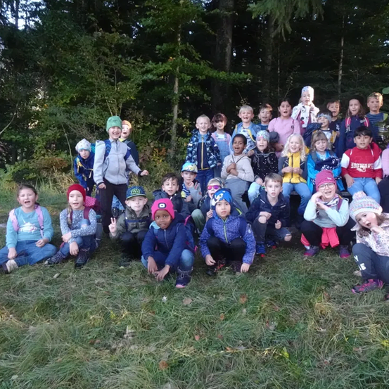 Eine Gruppe von Kindern und Erwachsenen posiert für ein Foto im Wald. Einige sitzen, andere stehen. Sie tragen Winterkleidung und manche haben Rucksäcke. Bäume und Gras umgeben sie.