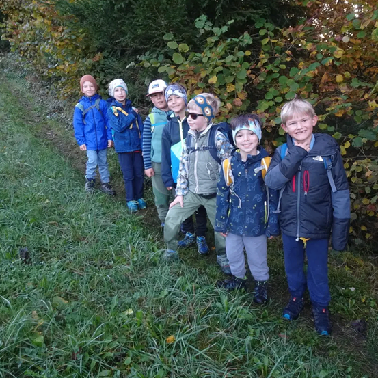 Eine Gruppe von Kindern in warmen Kleidungsstücken steht auf einem Grasweg, umgeben von herbstlichen Bäumen.