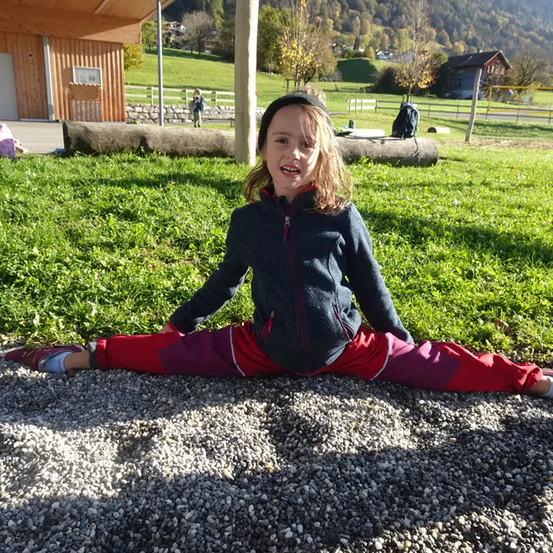 Ein kleines Mädchen in roten Hosen sitzt lächelnd auf Kies, mit einem Rasenbereich und Häusern im Hintergrund.