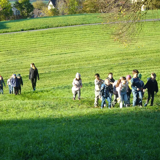 Eine Gruppe von Kindern und Erwachsenen geht auf einem Grasfeld, eine Frau ist in der Ferne zu sehen. Bäume und Häuser sind im Hintergrund sichtbar.