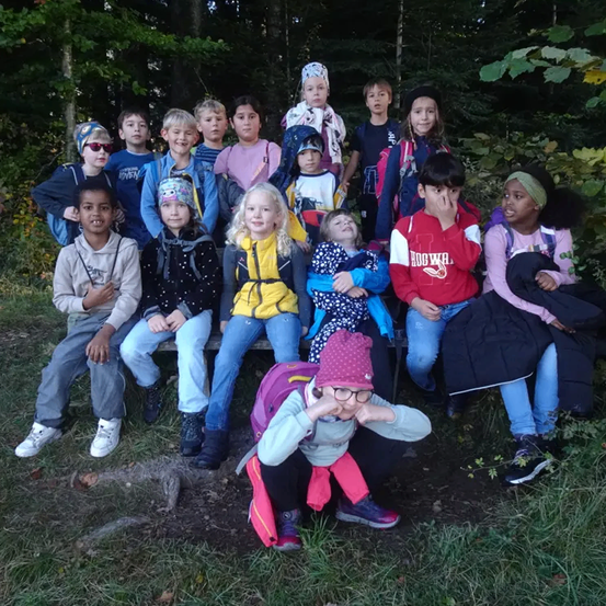 Eine Gruppe von Kindern sitzt auf einer Bank im Wald. Einige tragen Rucksäcke, andere Mützen. Das Mädchen vorne hockt, die anderen sitzen. Der Hintergrund besteht aus einer Mischung aus Bäumen und Gras.