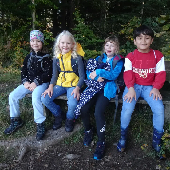 Vier Kinder sitzen auf einer Bank im Wald. Sie alle lächeln und scheinen auf einer Wanderung zu sein.