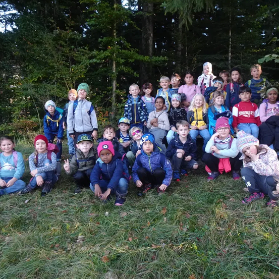 Eine Gruppe von Kindern posiert für ein Foto auf einem Grasfeld mit Bäumen im Hintergrund. Einige von ihnen sitzen, während andere stehen.