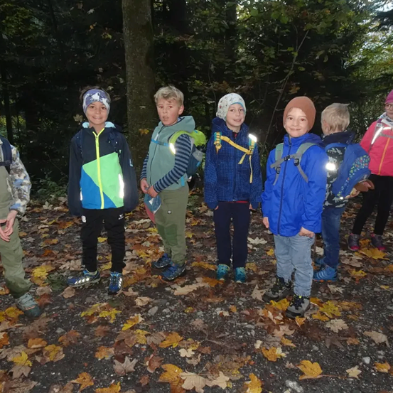 Eine Gruppe von Kindern steht auf einem Pfad im Wald und lächelt. Sie tragen Rucksäcke und einige haben reflektierende Westen. Der Boden ist mit Blättern bedeckt.