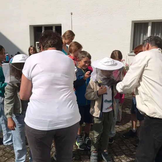 Eine Gruppe von Menschen, darunter Kinder, versammelt sich vor einem Gebäude. Einige tragen Imkerhauben, was auf eine Bildungs- oder Imkereiveranstaltung hindeutet.