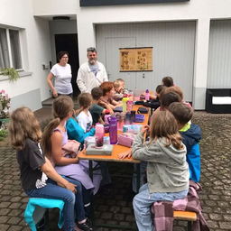 Eine Gruppe von Kindern sitzt um einen Tisch herum, mit einem Mann und einer Frau, die hinter ihnen stehen. Sie scheinen ein Picknick im Freien zu machen.