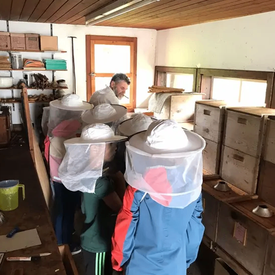 Eine Gruppe von Kindern steht in einem Raum und trägt Imkermützen. Ein Mann steht vor ihnen. Es gibt Holzkisten und einen Tisch mit einem Krug darauf.