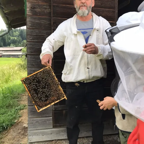 Ein Mann in einem Imkeranzug hält ein Bienenwabenstück vor einem Holzgebäude. Eine andere Person mit einem Gesichtsschutz steht in der Nähe.
