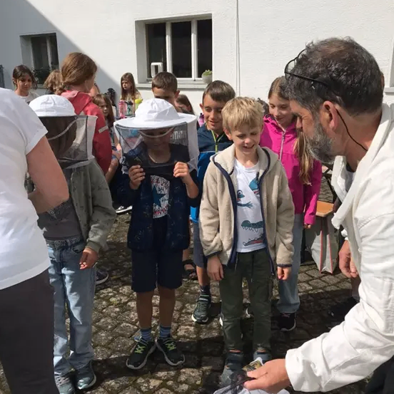 Eine Gruppe von Kindern und ein Mann in Schutzkleidung stehen im Freien. Sie halten ein Netz, möglicherweise um Insekten zu fangen. Der Mann trägt einen Imkerhut.