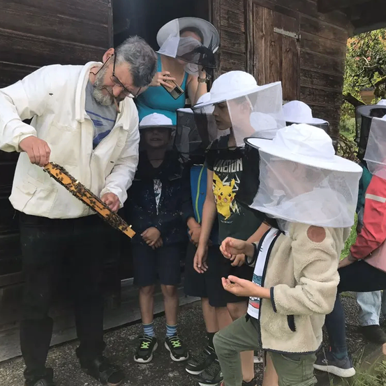 Ein erwachsener Mann, der einen Imkeranzug trägt, zeigt Kindern einen Bienenrahmen. Die Kinder, die Schutzschleier tragen, beobachten aufmerksam. Sie befinden sich in einem Holzschuppen.