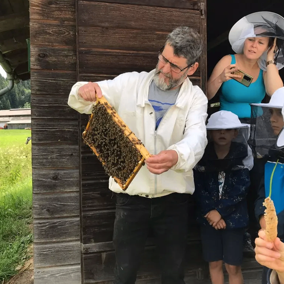 Ein Imker hält ein Bienenvolk in einer Holzhütte, während eine Frau und Kinder zusehen, einer hält einen Stock.