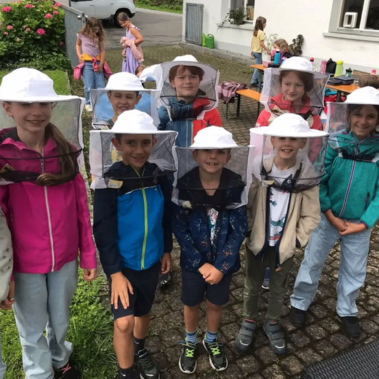 Eine Gruppe von Kindern in Bienenanzügen lächelt für die Kamera, während sie auf einem gepflasterten Boden mit Grünpflanzen und Gebäuden im Hintergrund stehen.