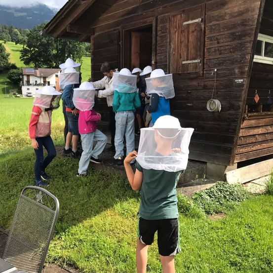 Eine Gruppe von Kindern in Imkerausrüstung steht vor einem Holzhaus auf einer Wiese. Sie lernen etwas über Bienen.