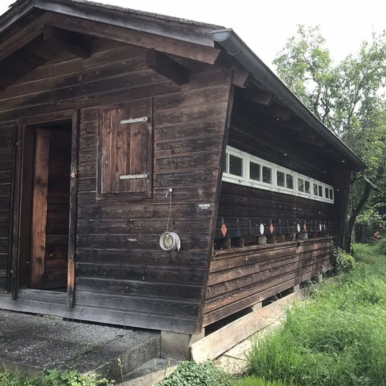 Ein verwittertes Holzhaus mit offener Tür, mehreren Fenstern und einem geneigten Dach. Ein hängendes weißes Objekt befindet sich in der Nähe der Tür.