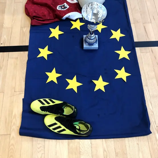 Ein Fußballtrikot, ein Fußball, ein Pokal und zwei Paar Schuhe liegen auf einem blauen Tuch mit gelben Sternen, möglicherweise zur Darstellung der Europäischen Union.