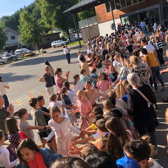 Eine Menschenmenge, bestehend aus Kindern und Erwachsenen, steht in einem Kreis vor einem Gebäude, wahrscheinlich einer Schule, an einem sonnigen Tag. Einige tanzen, während andere klatschen.