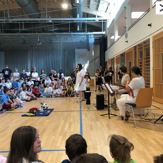 In einem Innenraum sind eine Gruppe von Menschen versammelt. Eine Frau singt in ein Mikrofon, während eine andere Frau Gitarre spielt. Das Publikum besteht aus Kindern und Erwachsenen.