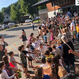 Eine große Menschenmenge, einschließlich Kinder, steht vor einem Gebäude. Sie bilden eine Schlange, manche halten Händchen, auf einem Parkplatz mit geparkten Autos.