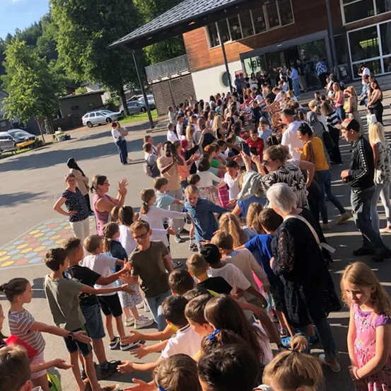 Eine große Menschenmenge aus Kindern und Erwachsenen steht in einer Reihe vor einem Gebäude. Einige winken und machen Fotos. Bäume und geparkte Autos sind im Hintergrund zu sehen.