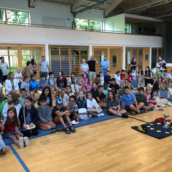 In einer Turnhalle sitzen Kinder auf blauen Matten und beobachten ein Ereignis. Ein Pokal und eine europäische Flagge liegen auf dem Boden. Zuschauer, darunter Erwachsene, beobachten hinter den Kindern.