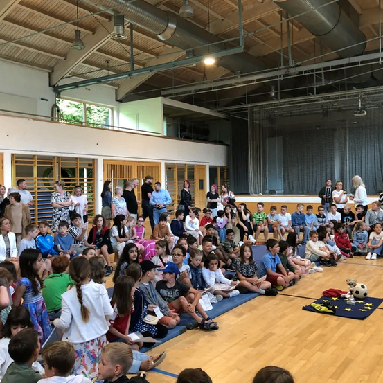 Eine große Menschenmenge aus Kindern und Erwachsenen versammelt sich in einer Turnhalle. Einige sitzen auf Matten, während andere stehen. Ein Pokal und ein Fußball liegen auf einer blauen Matte auf dem Boden.