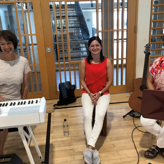 Drei Frauen sind drinnen und lächeln. Eine sitzt auf einem Hocker mit Gitarre und Notenständer. Eine andere steht neben einem Keyboard und die dritte neben einer Tür.