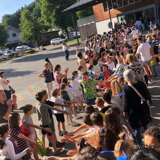 Eine große Gruppe von Kindern und Erwachsenen versammelt sich im Freien. Sie scheinen ein Ereignis zu feiern. Einige klatschen, und einige halten Händchen.