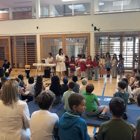 Eine Gruppe von Kindern und Erwachsenen versammelt sich in einem Raum, einige stehen und andere sitzen auf dem Boden. Eine Frau steht vor einem Klavier.