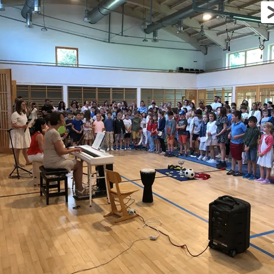 Eine Frau spielt Klavier, während eine große Menschenmenge aus Kindern und Erwachsenen zuschaut. Ein Schlagzeug und ein Fußball liegen auf dem Boden.