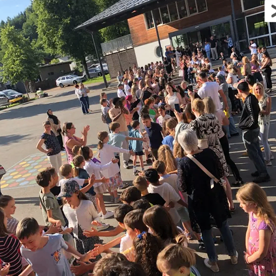 Eine Gruppe von Kindern und Erwachsenen steht in einer Reihe, wahrscheinlich für eine Veranstaltung, vor einem Gebäude mit vielen Fenstern.