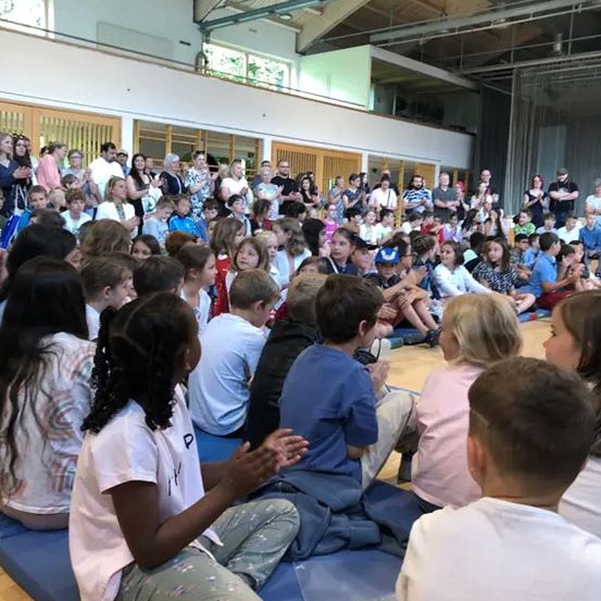 Eine große Gruppe von Kindern sitzt auf blauen Matten in einer Turnhalle und hört aufmerksam jemandem im Vordergrund zu. Im Hintergrund stehen und sitzen viele Erwachsene, einige klatschen.