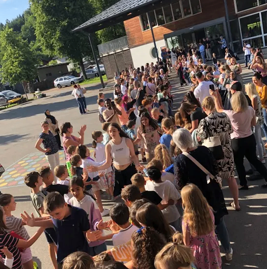 Eine große Menschenmenge aus Kindern und Erwachsenen versammelt sich an einem sonnigen Tag vor einem Gebäude, einige halten Händchen und lächeln, mit Bäumen und geparkten Autos im Hintergrund.