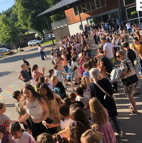 Eine Menschenmenge, bestehend aus Kindern und Erwachsenen, versammelt sich vor einem Gebäude mit einem Metalldach. Einige klatschen, während andere gehen. Im Hintergrund sind Bäume und geparkte Autos zu sehen.
