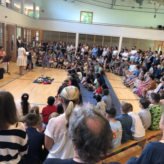 Eine große Gruppe von Menschen versammelt sich in einer Turnhalle. Einige sitzen auf Bänken, während andere stehen. Eine Frau steht auf dem Boden und hält möglicherweise eine Rede. Kinder sitzen auf dem Boden und ein Fußball liegt auf einem blauen Teppich.