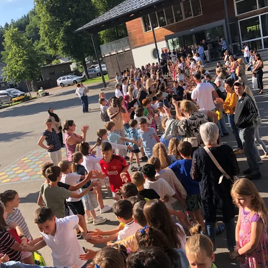 Eine Menschenmenge, einschließlich Kinder, Erwachsene und einige Senioren, versammelt sich vor einem Gebäude. Einige klatschen, während andere stehen und sich umsehen. Im Hintergrund sind Bäume, Autos und ein Weg sichtbar.