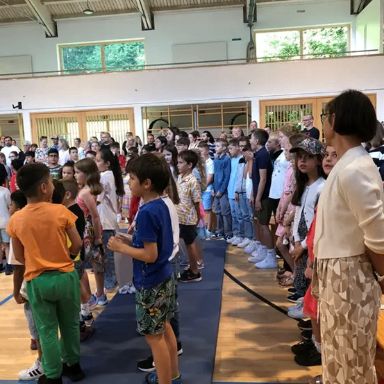 Eine Gruppe von Kindern und Erwachsenen steht in einer Reihe in einer Turnhalle. Einige Kinder tragen Mützen. Eine Frau steht vor ihnen.